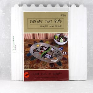 May include: A craft pattern titled "Threads That Bind" by designer Shawn Williams. The pattern features a colorful, oval-shaped applique design with a house, sun, and other elements. The pattern is in a clear plastic sleeve.