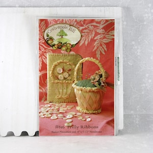 May include: A craft pattern titled "#816 Frilly Ribbons" featuring a basket pincushion and a 4" x 5 1/2" needlecase. The image shows a basket-shaped pincushion, a needlecase, and scattered buttons, all in soft, muted colors.