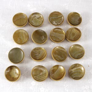 May include: A collection of sixteen round, natural stone cabinet knobs. Each knob features a unique pattern of tan, brown, and green hues, set within a gold-toned metal frame. The knobs are arranged in a grid pattern, showcasing their varied textures.