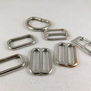 May include: A set of seven silver metal slider buckles, including two double-loop buckles, two single-loop buckles, a rectangular buckle, a D-ring, and a buckle with a center bar.
