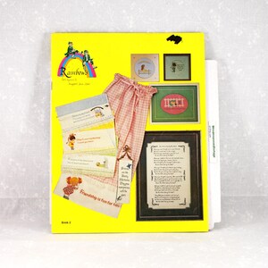 May include: A yellow book cover with the title "Rainbow's" and illustrations. The image includes cross-stitch samplers, a pair of pink gingham pants, and several embroidered cards with text. The phrase "Friendship is fun for two!" is visible.