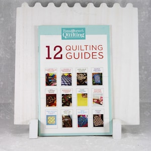 May include: A white wooden book stand holding a quilting guide. The guide is titled "Fons & Porter's Quilting" and features 12 quilting guides. The cover is light blue with red text and images of quilts.