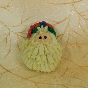 Peut inclure: Une petite figurine en c&eacute;ramique blanche de P&egrave;re No&euml;l avec un chapeau rouge, une garniture verte et une barbe blanche. La figurine a un style fantaisiste et cartoon.