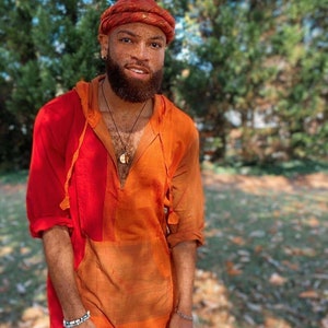 May include: A man wearing a red and orange hooded tunic with a red turban. He has a beard and is wearing a gold necklace with a pendant.