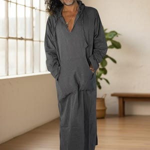 May include: A long, charcoal gray hooded robe with a V-neck and front pockets. The robe is floor-length and has long sleeves. The model is standing in a well-lit room with a large window and wooden floor.