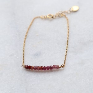 Op de afbeelding: Een delicate gouden kettingarmband met een rij kleine roze en rode edelstenen.