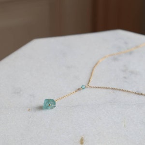 Könnte beinhalten: Eine zarte goldene Kettenkette mit einem kleinen, hellblauen Edelstein-Anhänger. Der Anhänger hat eine facettierte, unregelmäßige Form.