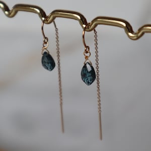 Peut inclure: Une paire de boucles d'oreilles pendantes en or avec une pierre précieuse bleu foncé sur chaque boucle d'oreille. Les boucles d'oreilles sont suspendues à un support en or.