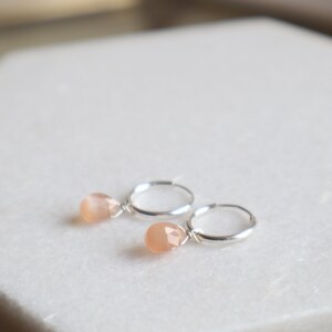 Op de afbeelding: Een paar zilveren oorringen met een klein, perzikkleurig edelsteentje dat aan elke oorring hangt.