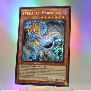Puede incluir: Una carta coleccionable de Yu-Gi-Oh! titulada Fydraulis Harmonia, con una ilustración holográfica de una criatura dragón. La carta tiene un borde marrón, ocho iconos de estrellas naranjas y texto que detalla los atributos de Dragón, Cantante y Efecto.
