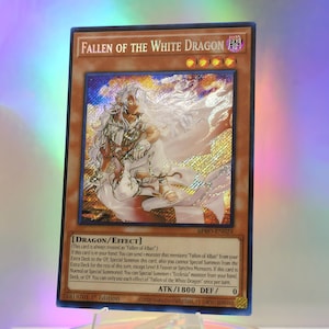 Peut inclure: Une carte à collectionner rectangulaire intitulée Fallen of the White Dragon avec une bordure brune et une illustration holographique d'un personnage aux cheveux blancs en robe blanche. La carte présente du texte, quatre étoiles et ATK 1800 DEF 0.
