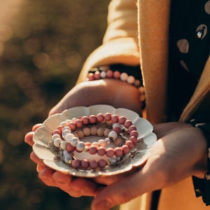 Könnte beinhalten: Drei rosafarbene, weiße und hölzerne Perlenarmbänder, die auf einer kleinen weißen Untertasse mit Blumenmuster gestapelt sind.