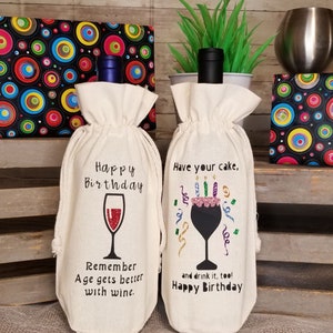 May include: Two white fabric wine bottle bags with birthday themed designs. The bag on the left has a wine glass with the text "Happy Birthday. Remember Age gets better with wine." The bag on the right has a wine glass with a cake on top and the text "Have your cake, and drink it too! Happy Birthday."