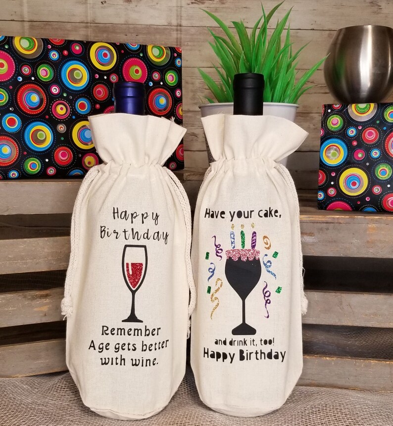May include: Two white fabric wine bottle bags with birthday themed designs. The bag on the left has a wine glass with the text "Happy Birthday. Remember Age gets better with wine." The bag on the right has a wine glass with a cake on top and the text "Have your cake, and drink it too! Happy Birthday."