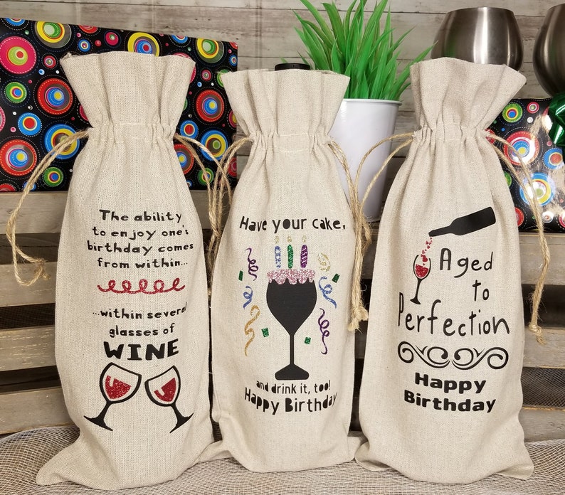 May include: Three beige linen wine bottle gift bags with different birthday sayings. The first bag says "The ability to enjoy one's birthday comes from within... within several glasses of WINE". The second bag says "Have your cake. And drink it too! Happy Birthday". The third bag says "Aged to perfection. Happy Birthday".