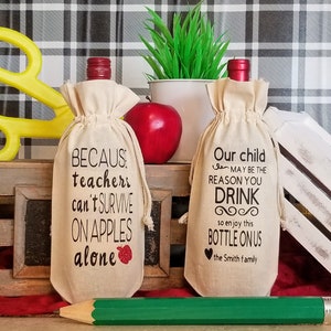 May include: Two beige wine bottle bags with black text. The bag on the left says "BECAUS: teachers can't SURVIVE ON APPLES alone" with a red apple graphic. The bag on the right says "Our child MAY BE THE REASON YOU DRINK so enjoy this BOTTLE ON US the Smith family".
