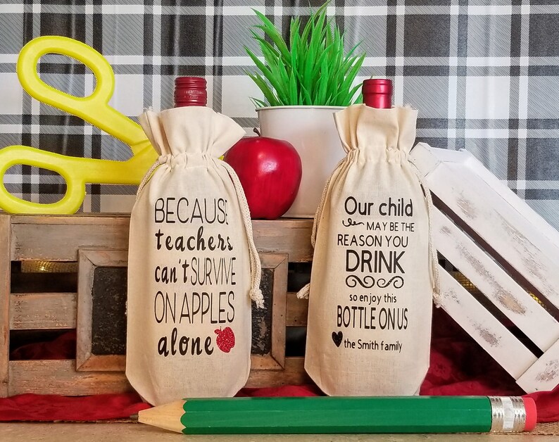 May include: Two beige wine bottle bags with black text. The bag on the left says "BECAUS: teachers can't SURVIVE ON APPLES alone" with a red apple graphic. The bag on the right says "Our child MAY BE THE REASON YOU DRINK so enjoy this BOTTLE ON US the Smith family".
