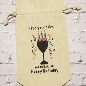 May include: A beige fabric gift bag with a drawstring closure. The bag has a black graphic of a wine glass with a cake on top and colorful candles. The text on the bag reads "Have your cake, and drink it, too! Happy Birthday."