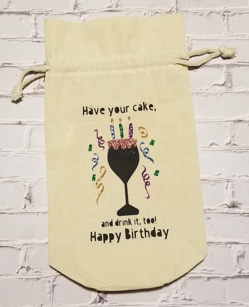 May include: A beige fabric gift bag with a drawstring closure. The bag has a black graphic of a wine glass with a cake on top and colorful candles. The text on the bag reads "Have your cake, and drink it, too! Happy Birthday."