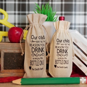 May include: Two burlap wine bottle bags with black text that reads "Our child may be the reason you drink so enjoy this bottle on us the Smith family".
