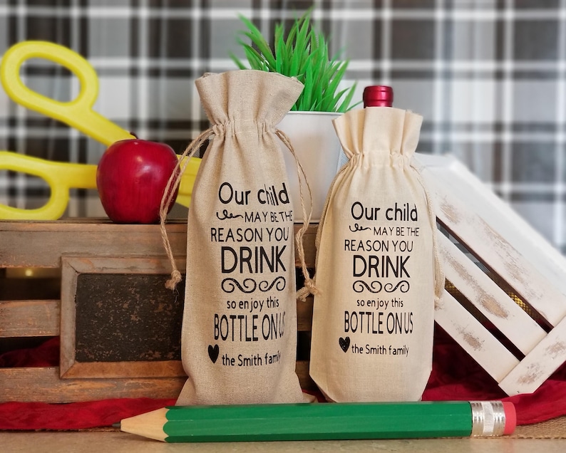 May include: Two burlap wine bottle bags with black text that reads "Our child may be the reason you drink so enjoy this bottle on us the Smith family".