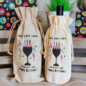 May include: Two beige wine bottle gift bags with a black silhouette of a wine glass with a pink cake and candles on top. The text on the bags reads "Have your cake, and drink it too! Happy Birthday".