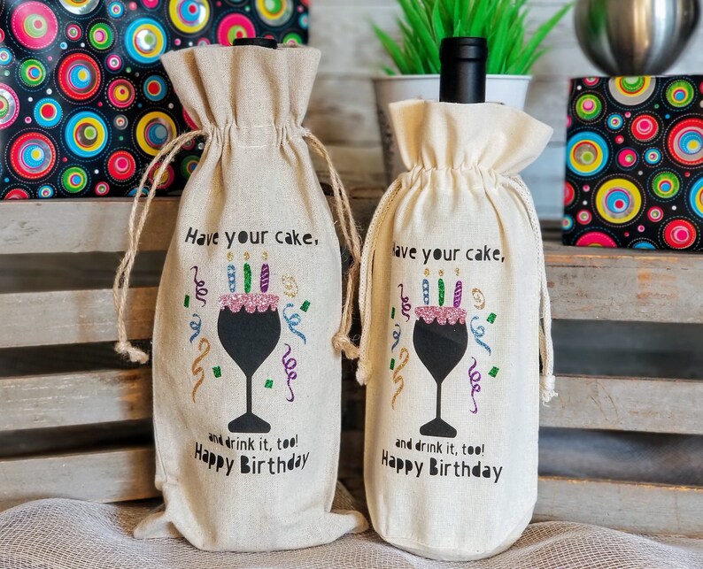 May include: Two beige wine bottle gift bags with a black silhouette of a wine glass with a pink cake and candles on top. The text on the bags reads "Have your cake, and drink it too! Happy Birthday".