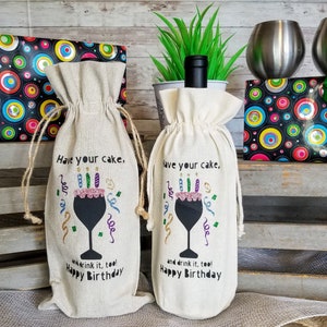 May include: Two white fabric wine bags with a black wine glass design and the text "Have your cake, and drink it, too! Happy Birthday".