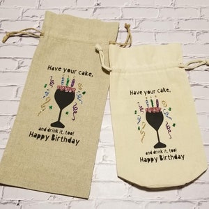 May include: Two beige fabric wine bottle gift bags with a black silhouette of a wine glass with a cake and candles inside. The text on the bags reads "Have your cake, and drink it, too! Happy Birthday".