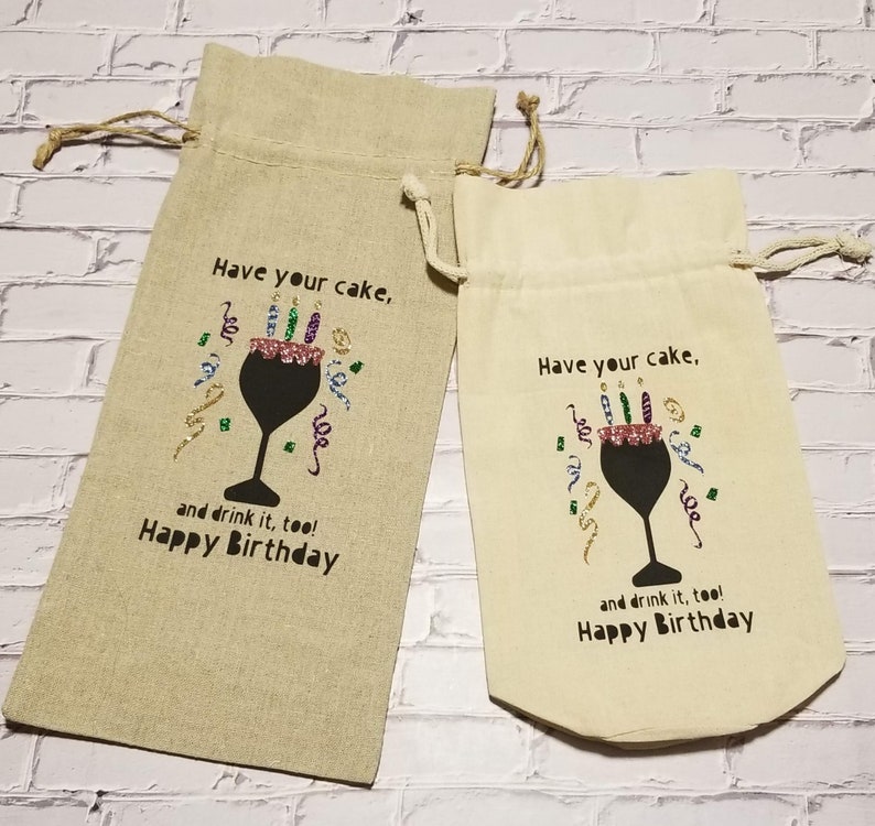 May include: Two beige fabric wine bottle gift bags with a black silhouette of a wine glass with a cake and candles inside. The text on the bags reads "Have your cake, and drink it, too! Happy Birthday".