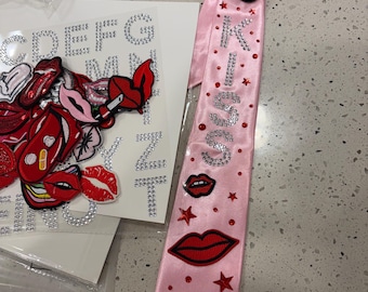 Op de afbeelding: Een lichtroze satijnen stropdas versierd met rode geborduurde lippenpatches, kleine rode sterren en het woord KISS gespeld in zilveren strass-steentjes. De das wordt getoond naast een vel met opstrijkbare lippenpatches en alfabetletters.
