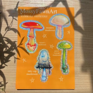 May include: An orange sticker featuring four colorful mushroom illustrations with glittery outlines. The sticker includes the text "MossyFawnArt technicolor mushrooms" and the names of each mushroom: caesar's mushroom, meadow mushroom, inky cap mushroom, and parrot waxcap mushroom.