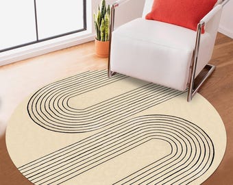Pode incluir: Um tapete redondo de cor creme com um design geométrico preto de linhas curvas e listras. O tapete está colocado sobre um piso de madeira na frente de uma cadeira branca com uma almofada vermelha.