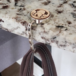 May include: A silver-toned hook with a wooden knob featuring a laser-engraved design of a snowman. The hook is attached to a brown canvas strap.