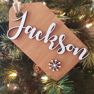 May include: A wooden Christmas ornament with the name "Jackson" written in white cursive lettering. A small white snowflake is in the bottom right corner of the ornament.