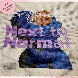 以下が含まれることがあります： 薄いベージュ色の生地に紫色の「Next to Normal」の文字がクロスステッチされたデザイン。青と黒の色合いのシルエットと、茶色と白のアクセントが特徴です。左上隅のピンク色の円には「PDF Cross Stitch Pattern」と書かれています。