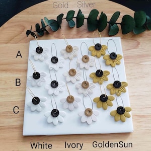 May include: A set of 12 pairs of handmade polymer clay earrings featuring a daisy flower design. The earrings are available in three color combinations: white with black centers, ivory with gold centers, and golden yellow with black centers. The earrings are labeled A, B, and C, and the colors are labeled Gold, Silver, White, Ivory, and GoldenSun.