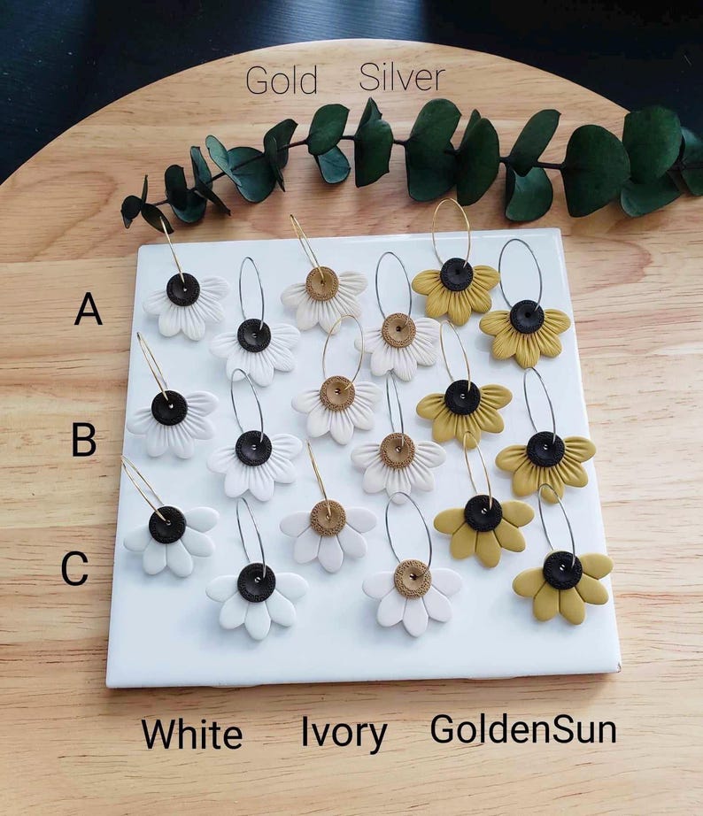 May include: A set of 12 pairs of handmade polymer clay earrings featuring a daisy flower design. The earrings are available in three color combinations: white with black centers, ivory with gold centers, and golden yellow with black centers. The earrings are labeled A, B, and C, and the colors are labeled Gold, Silver, White, Ivory, and GoldenSun.