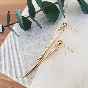 May include: A pair of gold dangle earrings with a long, thin bar and a small ball at the end. The earrings are on a white and brown surface.