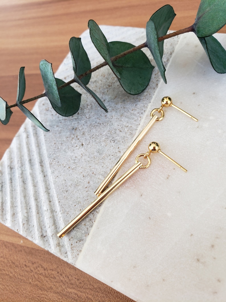 May include: A pair of gold dangle earrings with a long, thin bar and a small ball at the end. The earrings are on a white and brown surface.