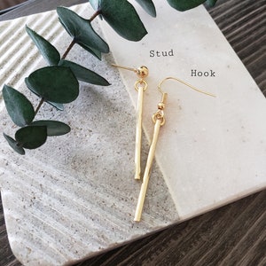 May include: A pair of gold earrings, one with a stud and the other with a hook. The stud earring has a small ball at the top and a long, straight bar. The hook earring has a small ball at the top and a curved hook.