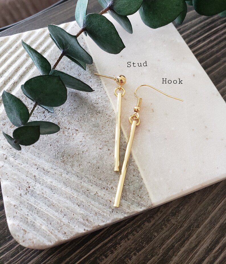 May include: A pair of gold earrings, one with a stud and the other with a hook. The stud earring has a small ball at the top and a long, straight bar. The hook earring has a small ball at the top and a curved hook.