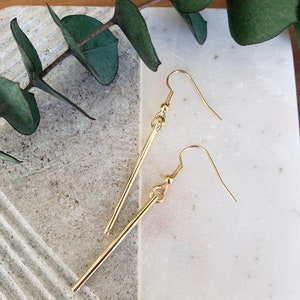 May include: A pair of gold-toned earrings with long, straight, thin bars hanging from small hoops. The earrings are on a light gray and white textured surface.