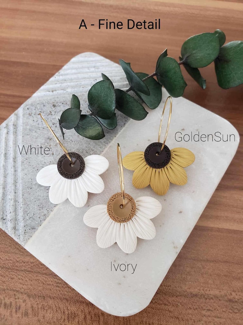 May include: Three pairs of flower-shaped earrings with gold hoops. The earrings are made of polymer clay and have a brown center with a gold accent. The flowers are white, ivory, and yellow.