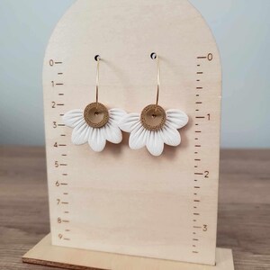 May include: A pair of white flower-shaped earrings with gold hoops and brown centers. The earrings are displayed on a wooden stand with a ruler.
