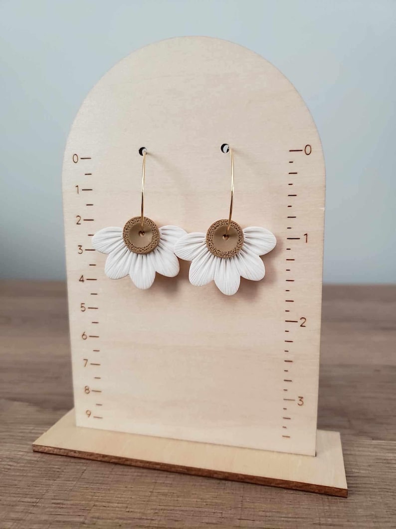May include: A pair of white flower-shaped earrings with gold hoops and brown centers. The earrings are displayed on a wooden stand with a ruler.