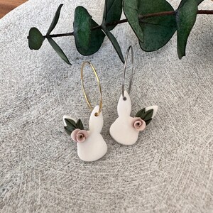 May include: A pair of white bunny-shaped earrings with pink flowers and green leaves. The earrings are on a white background.