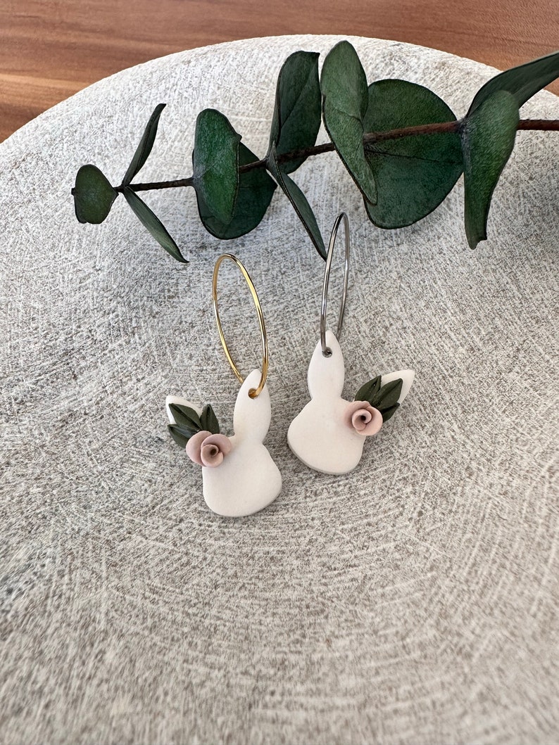 May include: A pair of white bunny-shaped earrings with pink flowers and green leaves. The earrings are on a white background.