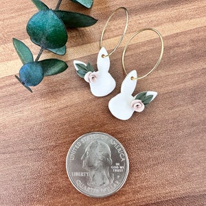 May include: A pair of white bunny-shaped earrings with pink flowers and green leaves hanging from gold hoops. The earrings are on a wooden surface.