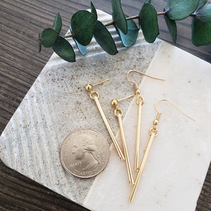 May include: Three pairs of gold bar earrings with a simple design. The earrings are on a white marble surface with a green plant branch.
